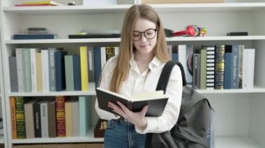 Young blonde woman student standing reading book at university classroom
