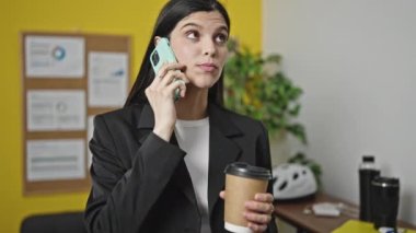 Young beautiful hispanic woman business worker talking on smartphone drinking coffee at office