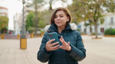 Orta yaşlı bir kadın, parktaki akıllı telefondan özçekim yaparken kendinden emin bir şekilde gülümsüyor.
