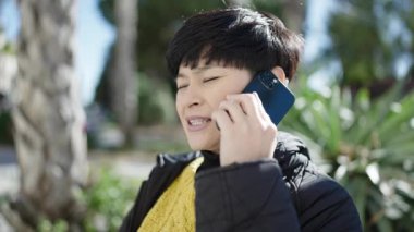 Genç Çinli kadın gülümsüyor. Parkta akıllı telefondan konuşuyor.