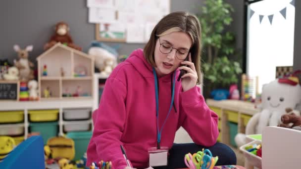 Young Blonde Woman Preschool Teacher Writing Document Talking Smartphone Kindergarten — Vídeo de Stock