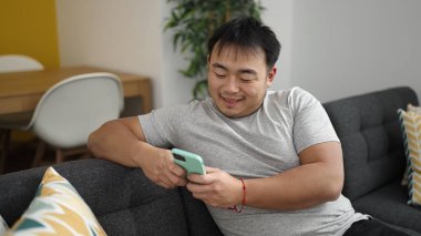 Young chinese man using smartphone sitting on sofa at home