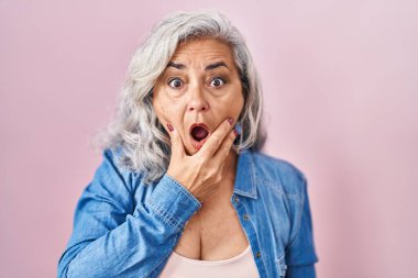 Middle age woman with grey hair standing over pink background looking fascinated with disbelief, surprise and amazed expression with hands on chin 