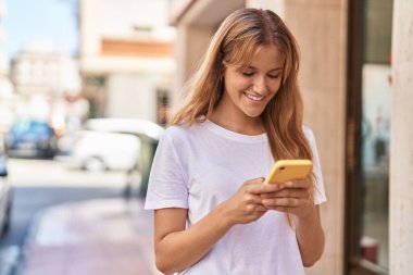 Young blonde girl smiling confident using smartphone at street