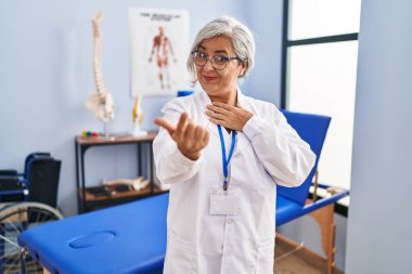 Gri saçlı orta yaşlı bir kadın ağrı kesici kliniğinde çalışıyor. El kol hareketi yaparak mutlu ve gülümseyen insanları davet ediyor. 