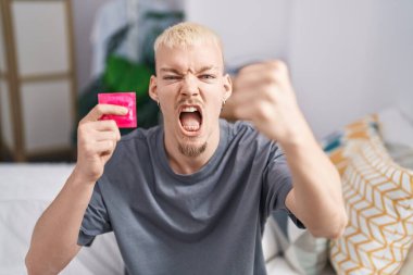 Kafkasyalı genç bir adam elinde prezervatifle yatakta sinirli ve sinirli bir şekilde oturmuş öfkeyle bağırıyor ve el kaldırıyor. 