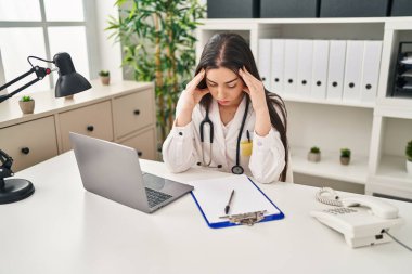 Young beautiful hispanic woman doctor stressed using laptop working at clinic