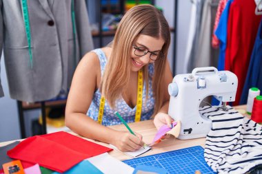Young beautiful hispanic woman tailor writing on notebook holding cloth at tailor shop