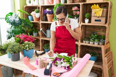 Young beautiful hispanic woman florist talking on smartphone writing on notebook at flower shop