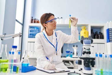 Laboratuvarda test tüpü tutan ve panoya yazı yazan bilim adamı üniforması giyen İspanyol genç bir kadın.