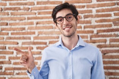 Tuğla duvarın üzerinde neşeli bir şekilde duran genç İspanyol adam yüzünde mutlu ve doğal bir ifadeyle elleri ve parmaklarıyla işaret eden bir gülümsemeyle. 