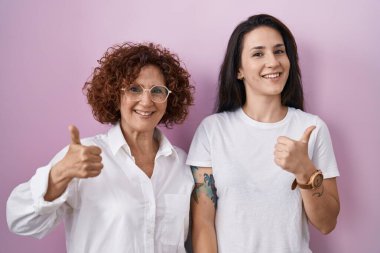 Hispanic mother and daughter wearing casual white t shirt over pink background doing happy thumbs up gesture with hand. approving expression looking at the camera showing success. 