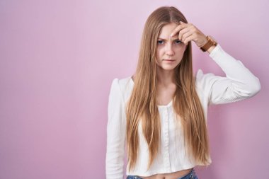 Young caucasian woman standing over pink background pointing unhappy to pimple on forehead, ugly infection of blackhead. acne and skin problem 