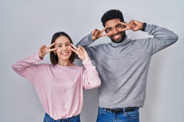 Young hispanic couple standing together doing peace symbol with fingers over face, smiling cheerful showing victory 
