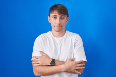 Kafkasyalı sarışın adam mavi arka planda duruyor. Kış soğuğu için titriyor ve donuyor. Yüzünde üzgün ve şok ifadesi var. 