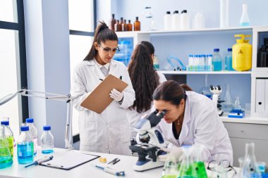 Three woman scientists using microscope write on checklist at laboratory