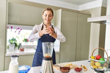 Genç sarışın kadın mutfaktaki blendere muz dökerek gülümsüyor.