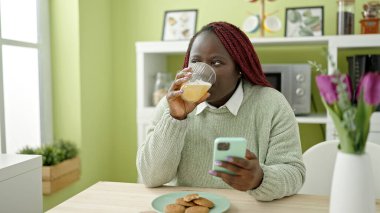 Örgülü saçlı Afrikalı kadın yemek odasında akıllı telefon kullanarak meyve suyu içiyor.