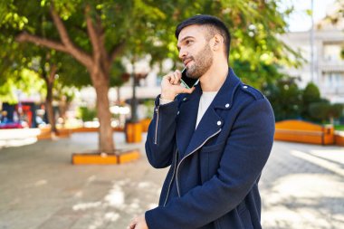 Young hispanic man talking on smartphone with serious expression at park
