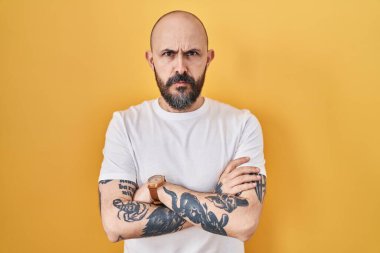 Sarı arka planda duran İspanyol dövmeli genç adam şüpheci ve gergin, yüzünde çapraz kollu tasvip etmeyen ifade. negatif kişi. 
