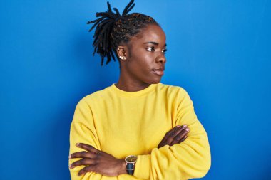 Beautiful black woman standing over blue background looking to the side with arms crossed convinced and confident 