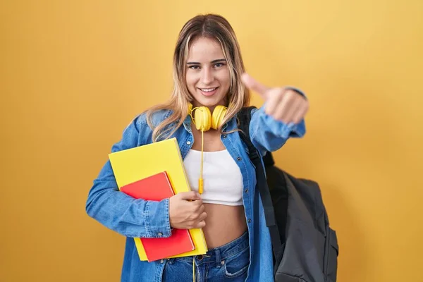 若いブロンドの女性は 学生のバックパックを着て 手で肯定的なジェスチャーを行うことを承認本を保持します 親指アップ笑顔と成功のために幸せ 勝者のジェスチャー — ストック写真