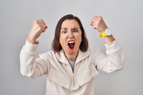 Middle age hispanic woman standing over isolated background angry and mad raising fists frustrated and furious while shouting with anger. rage and aggressive concept. 