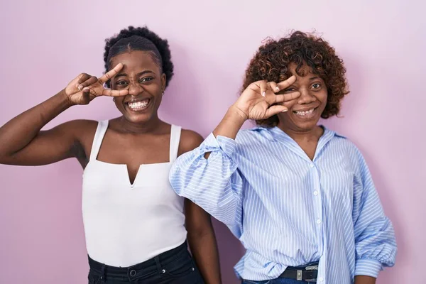 两个非洲女人站在粉红的背景上 用手指捂住脸 愉快地微笑着 表示胜利 — 图库照片