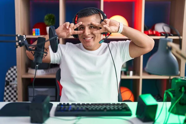 若いヒスパニック系の男が顔の上に指で平和のシンボルをやってビデオゲームをプレイし 明るい勝利を示す笑みを浮かべて — ストック写真