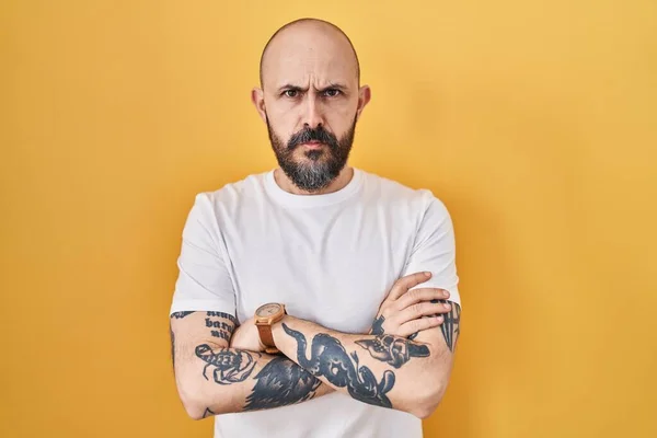 Young hispanic man with tattoos standing over yellow background skeptic and nervous, disapproving expression on face with crossed arms. negative person.