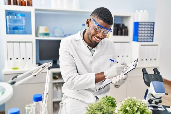 若いアフリカ系アメリカ人の男を着て科学者の制服を着てクリップボード上の書き込み植物で研究室 — ストック写真