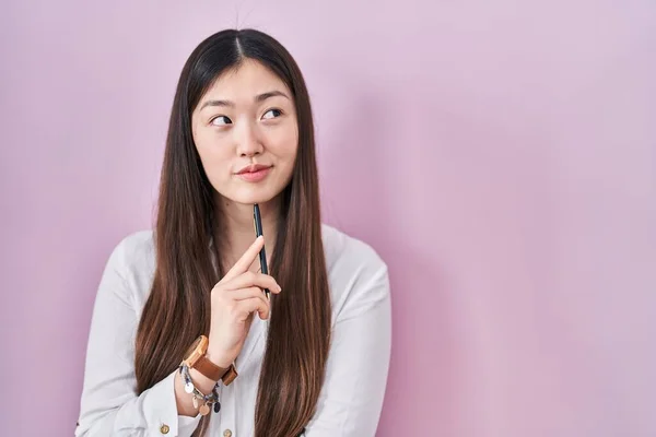 中国的年轻女子把铅笔放在粉色的背景上 面带微笑地看着旁边 目不转睛地看着外面想 — 图库照片