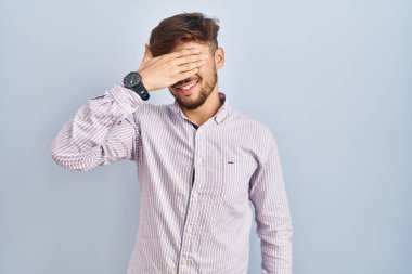 Mavi arka planda dikilen sakallı bir Arap. Gülümsüyor ve gülümsüyor. Gözlerini şaşkınlıkla kapatıyor. kör kavram. 