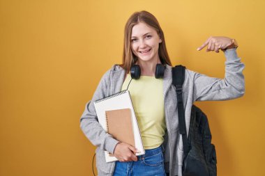 Sırt çantası takan, elinde kitaplarla gülümseyen, kendini gururlu ve mutlu gösteren genç beyaz bir kadın.. 