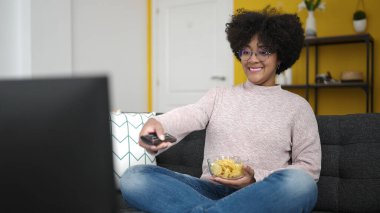 Genç Afrikalı Amerikalı kadın televizyon seyrediyor. Kanepede oturup cips yiyor.
