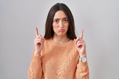 Genç esmer kadın beyaz arka planda dikiliyor üzgün ve üzgün görünüyor, parmaklarıyla yön gösteriyor, mutsuz ve depresif.. 