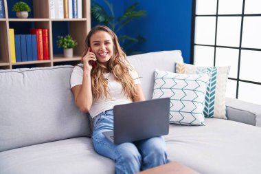Genç, güzel, İspanyol bir kadın akıllı telefonuyla evde dizüstü bilgisayar kullanıyor.