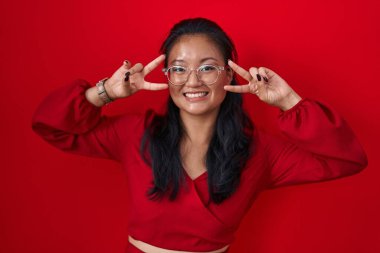 Asian young woman standing over red background doing peace symbol with fingers over face, smiling cheerful showing victory 