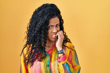 Middle age hispanic woman standing over yellow background looking stressed and nervous with hands on mouth biting nails. anxiety problem. 