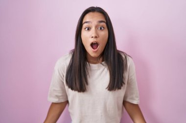 Young hispanic woman standing over pink background afraid and shocked with surprise and amazed expression, fear and excited face. 