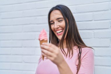 Young beautiful hispanic woman smiling confident eating ice cream over isolated white brick background