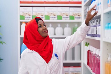 Young beautiful woman pharmacist smiling confident holding medication on shelving at pharmacy