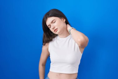 Young caucasian woman standing over blue background suffering of neck ache injury, touching neck with hand, muscular pain 