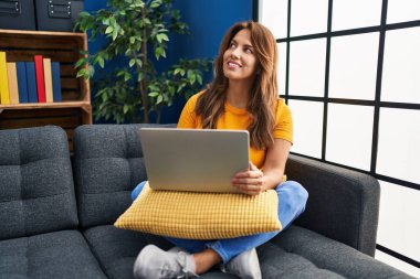 Evdeki kanepede dizüstü bilgisayar kullanan genç Latin kadın.