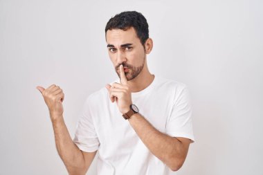 Beyaz arka planda dikilen yakışıklı İspanyol adam sessiz olmak istiyor. Parmakları dudaklarında, elleri yandan işaret ederek. sessizlik ve gizli konsept. 