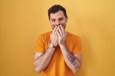 Young hispanic man standing over yellow background laughing and embarrassed giggle covering mouth with hands, gossip and scandal concept 