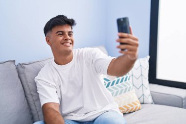 Genç ispanyol adam evdeki kanepede oturarak akıllı telefonuyla selfie çekiyor.
