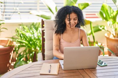 Young hispanic woman using laptop sitting on table at home terrace