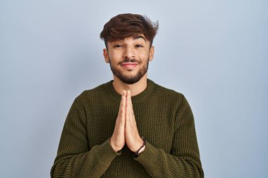 Arab man with beard standing over blue background praying with hands together asking for forgiveness smiling confident. 