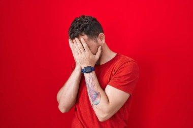 Genç İspanyol adam kırmızı arka planda duruyor. Ağlarken suratını ellerle kapatıyor. depresyon kavramı. 
